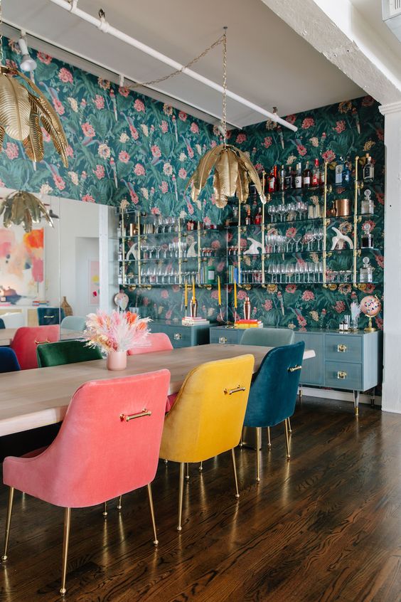 a bold dining room with a mirror wall, a blue home bar, glass shelves, a table, colorful matching chairs, chic gold pendant lamps