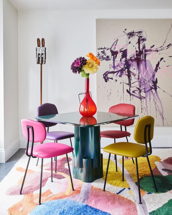 a bold dining space with a teal table, colorful chairs, a bold rug, a vase with bright blooms and an abstract artwork