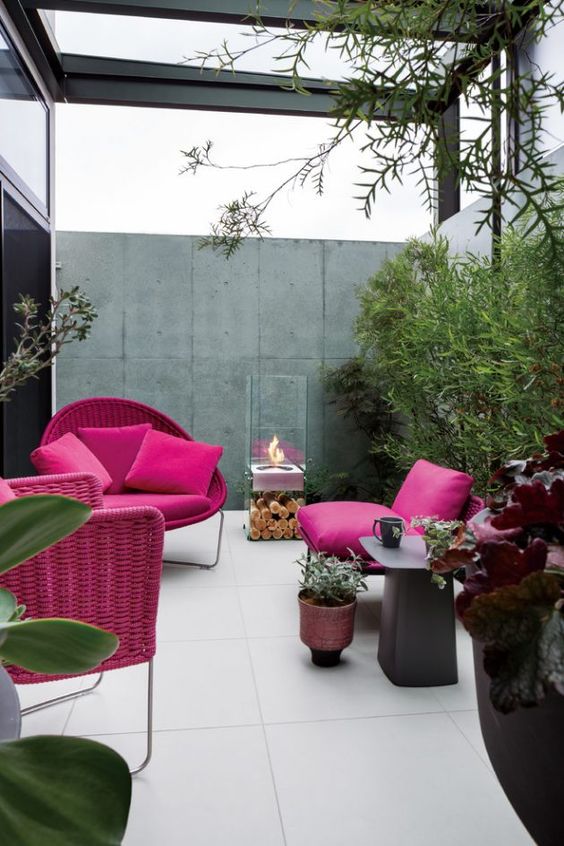a bold modern terrace withfuchsia furniture and pillows, greenery, a side table and a portable fireplace is cool