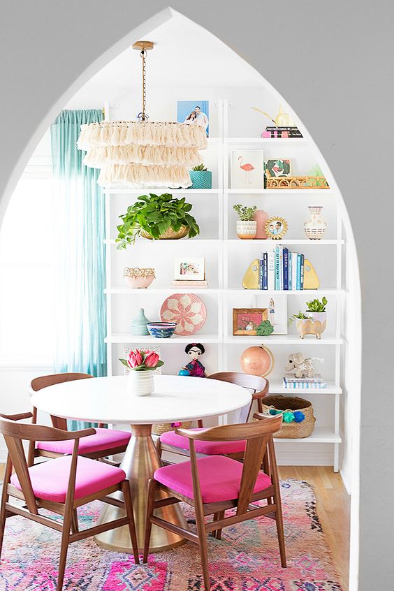 a bright and catchy dining space with hot pink chairs and a rug, a shelving unit with bright decor and aqua curtains