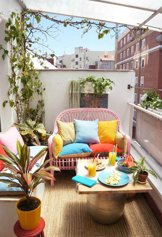 a bright balcony with a pink loveseat and a sofa, colorful pillows and bright tableware plus greenery around