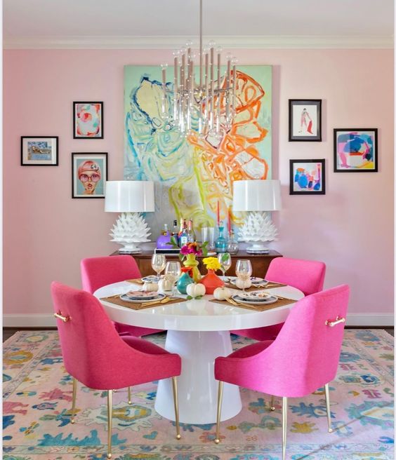 a bright dining room with blush walls, a round table, hot pink chairs, colorful artwork and a chic tube chandelier