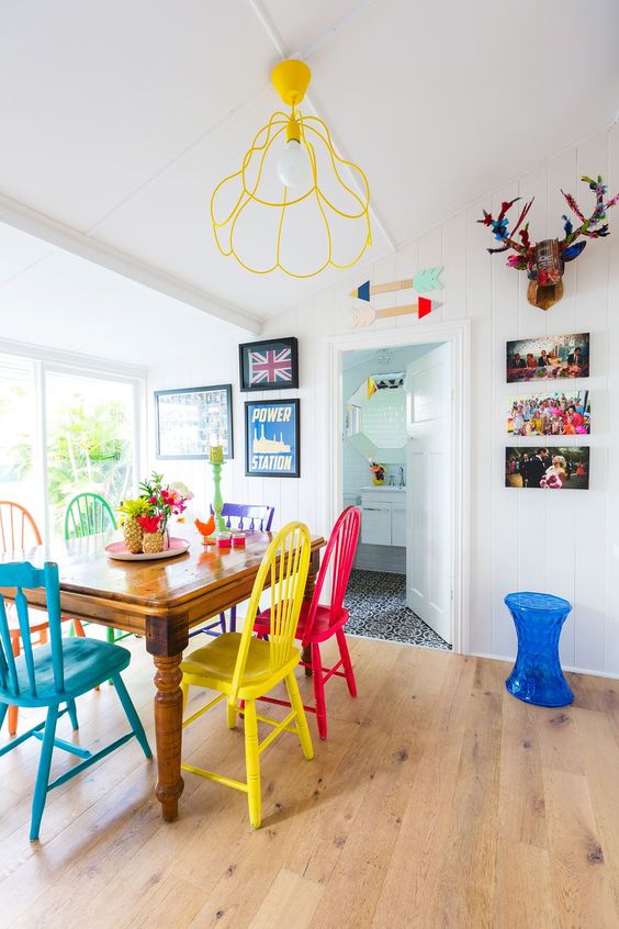 a bright eclectic dining room with a vintage table, colorful mismatching chairs, a yellow lamp, bright artwork and a side table