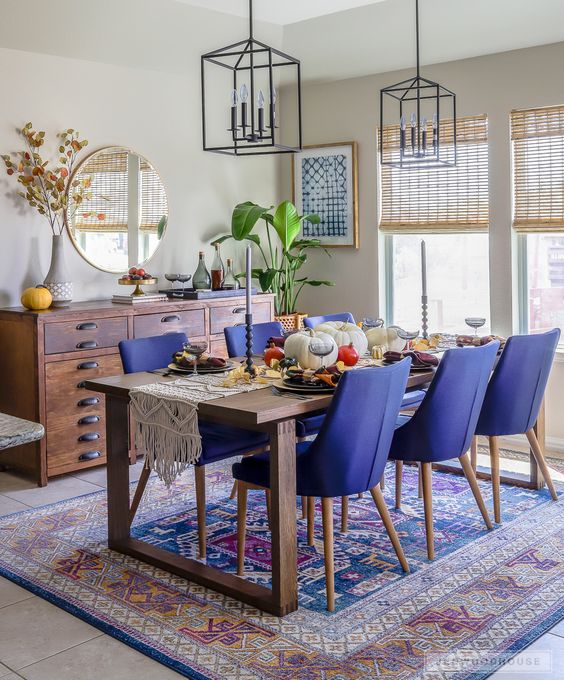 a bright farmhouse dining room with a blue and purple rug and cobalt chairs, a stained table and filed cabinet, pendant lamps