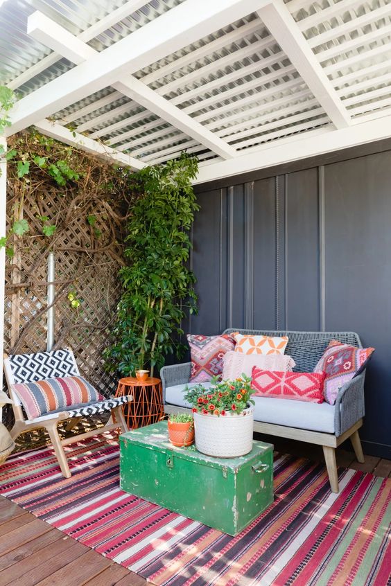 a bright terrace with a colorful striped rug, some furniture and bright pillows, a green chest with blooms and greenery