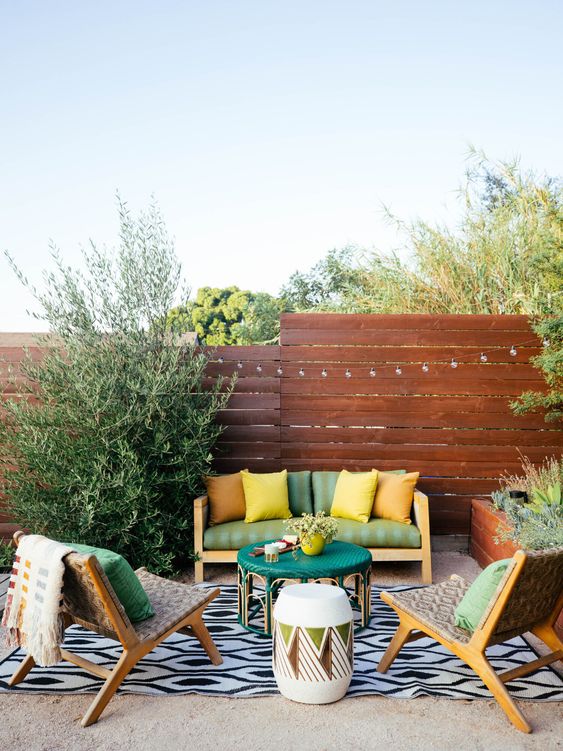 a bright terrace with green furniture, colorful pillows, a printed rug, an emerald table and lots of greenery around