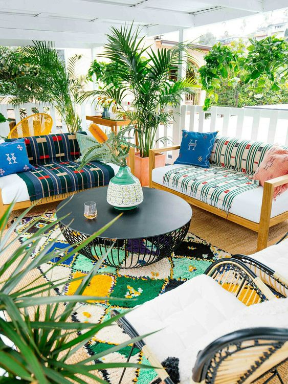 a bright terrace with sofas and printed blankets and pillows, a bright rug, a coffee table and potted plants and trees
