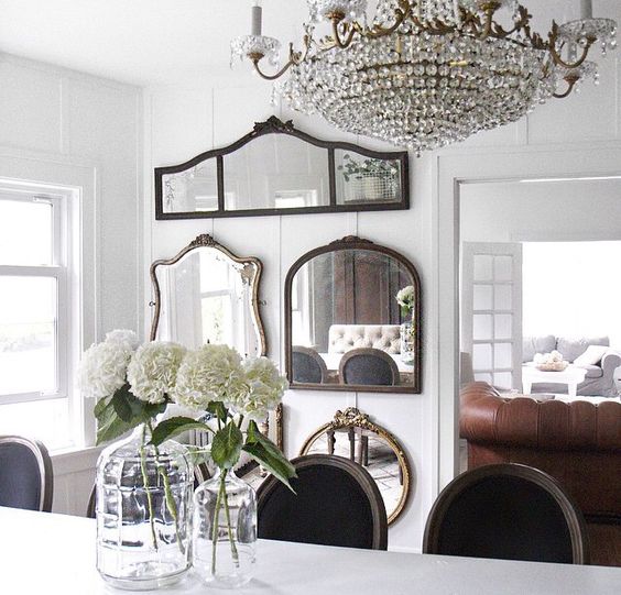 a chic dining room with a vintage mirror gallery wall, a white table, black chairs, a crystal chandelier and some blooms