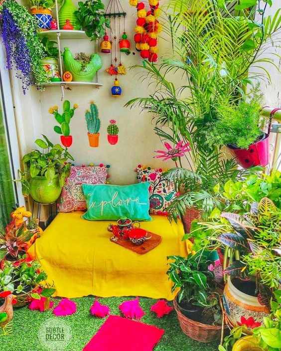 a colorful balcony with a green rug, a yellow daybed with colorful pillows, potted plants and blooms and pompoms and pillows