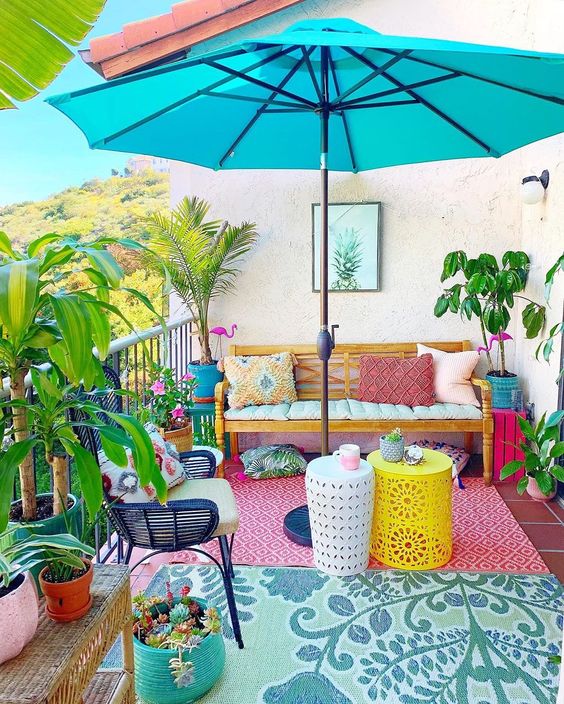 a colorful balcony with a loveseat and pillows, colorful rugs, side tables, lots of potted plants and a turquoise umbrella