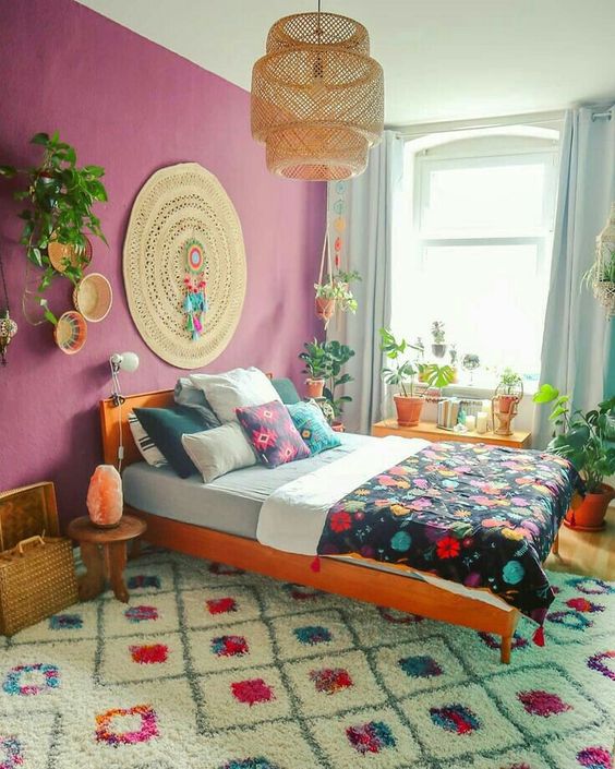 a colorful boho room with a purple wall, a jute rug, an orange bed with colorful bedding, a bold printed rug and potted plants
