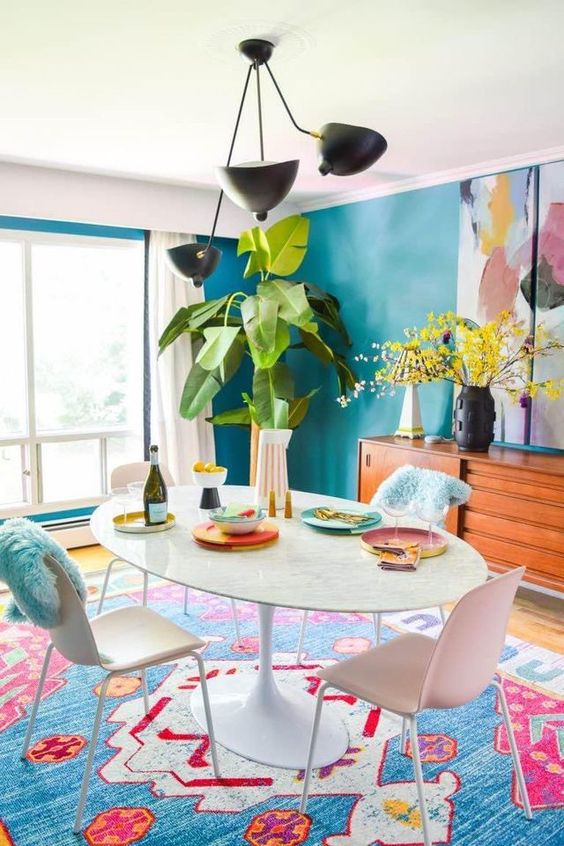 a colorful dining with blue walls, a colorful rug, a white table and chairs, greenery and a bold artwork