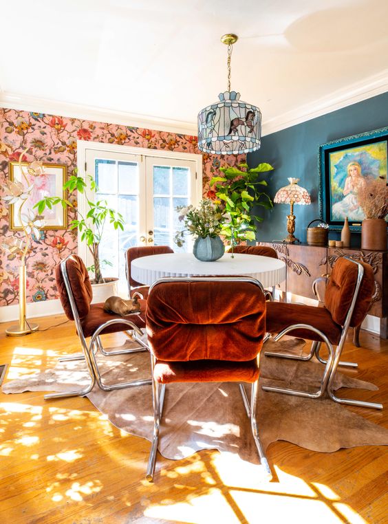 a colorful dining room with a navy and floral wall, a table, rust-colored chairs, a mosaic chandelier and some greenery