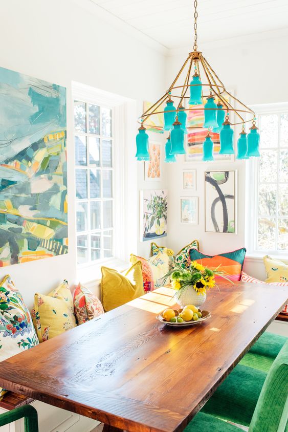 a colorful dining room with a stained table, green chairs, colorful pillows, a chandelier with turquoise lampshades and bold art