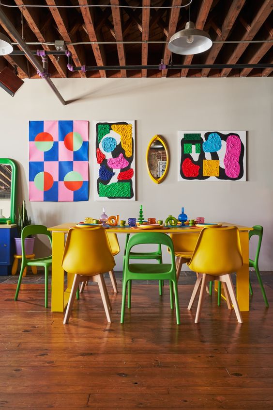 a colorful dining room with a yellow table, yellow and green chairs, colorful artwork, a mirror and some bright decor on the table