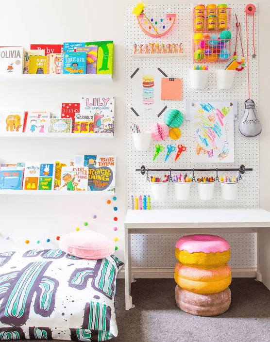 a colorful kids' room with a white desk, a pegboard with colorful decor and stuff, a bookshelf with books and a donut stool