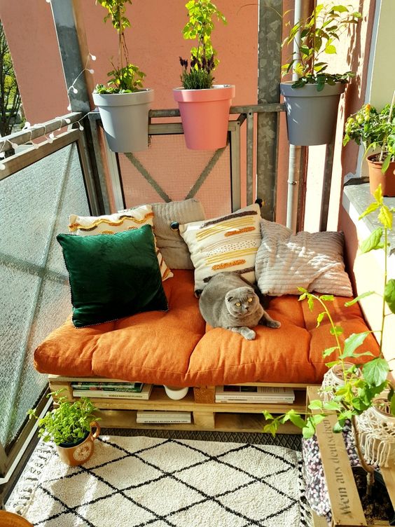 a colorful small balcony with a pallet sofa and bright pillows and books, potted greenery and blooms, with railing planters