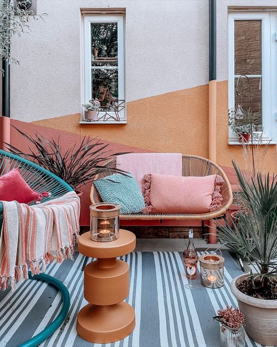 a colorful terrace with a color block wall, rattan furniture, a striped rug, a teal chair and bright pillows