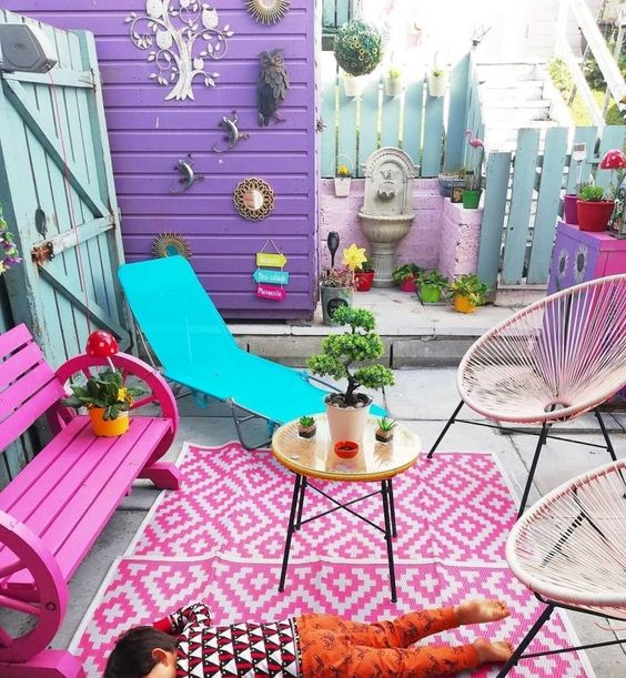 a colorful terrace with a purple wall, a hot pink bench, a bold rug, a turquoise lounger and some bright decor