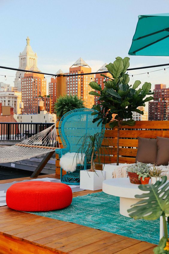 a colorful terrace with a turquoise papasan chair and a rug, a red pouf, some white furniture and potted plants and trees