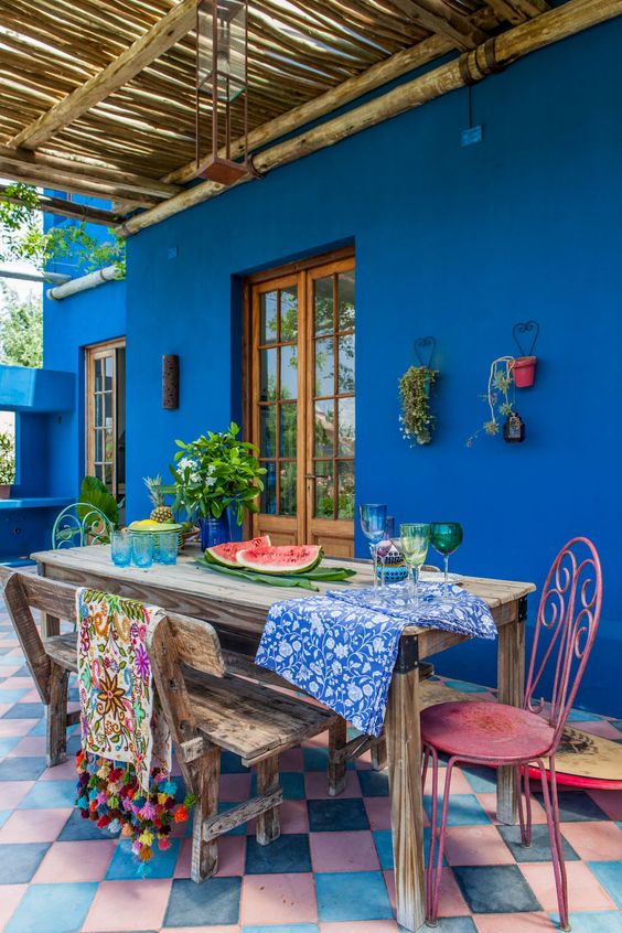 a colorful terrace with navy walls, a stained table and a bench, colorful chairs, colorful textiles and a bright tiled floor