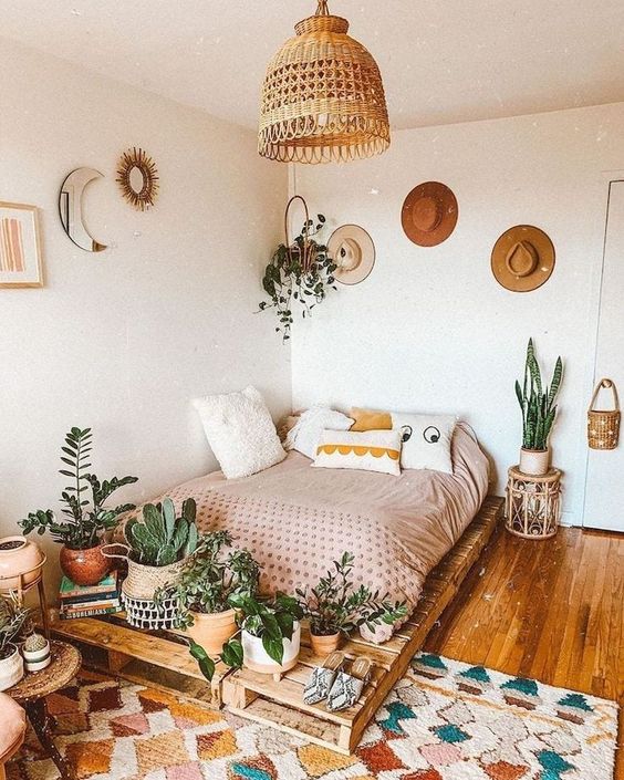 a cool boho bedroom with a pallet bed and neutral bedding, a colorful rug, a lot of plants, some boho decor and hats on the wall