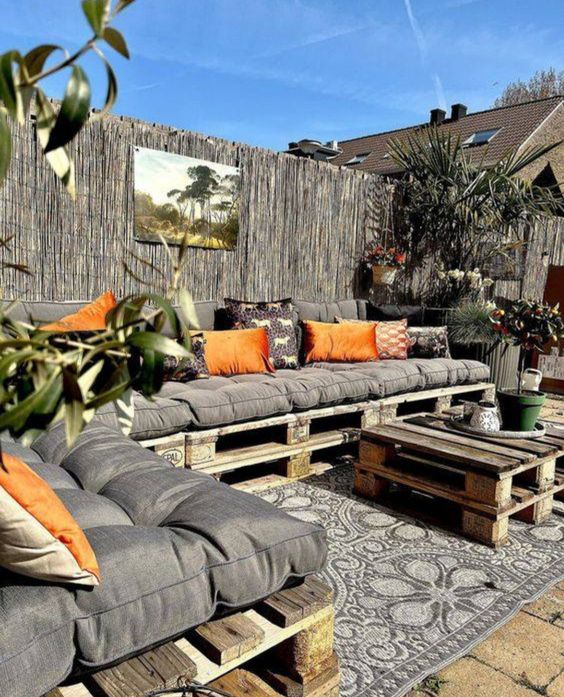a cool terrace with a large pallet sofa and grey cushions and orange pillows, a pallet table and lots of potted greenery is wow