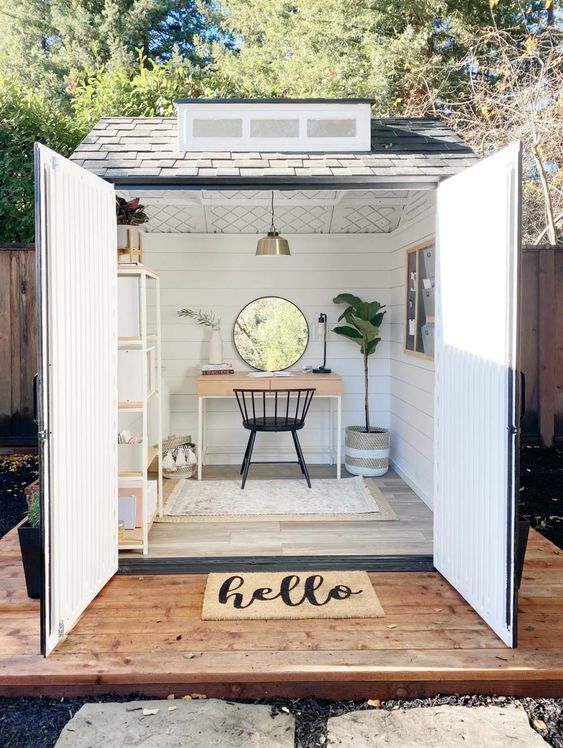 a cute shed home office with white planked walls, a desk, a shelving unit, a potted plant and a round window