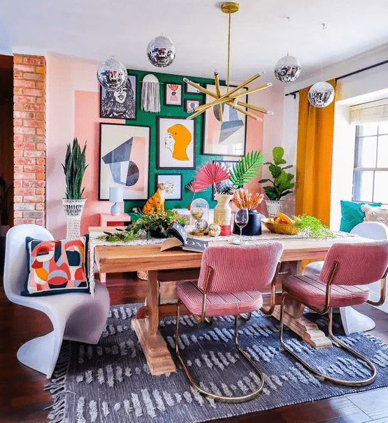 a dining room with a green accent wall and a bold gallery wall, a table, pink chairs, colorful pillows and greenery