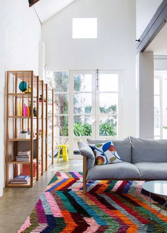 a double-height living room with a grey sofa and a colorful modern rug, a shelving unit with bright decor and a coffee table