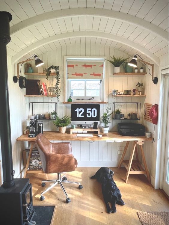 a farmhouse shed home office with a stained trestle desk, a leather chair, shelves with decor and potted plants is lovely