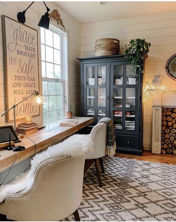 A farmhouse shed home office with a wall mounted desk, white chairs, a boho rug, a fireplace, a black storage unit, potted plants and artwork