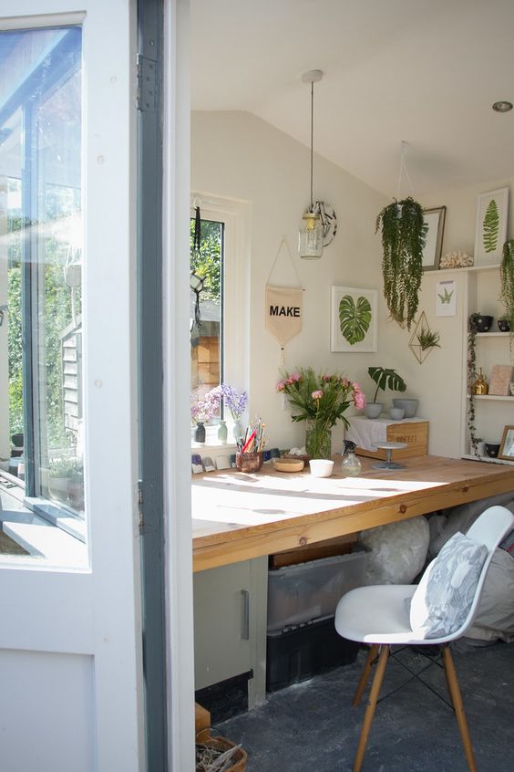 a garden home office with a large desk, open shelves, potted plants, artwork and a white chair is cozy and cute
