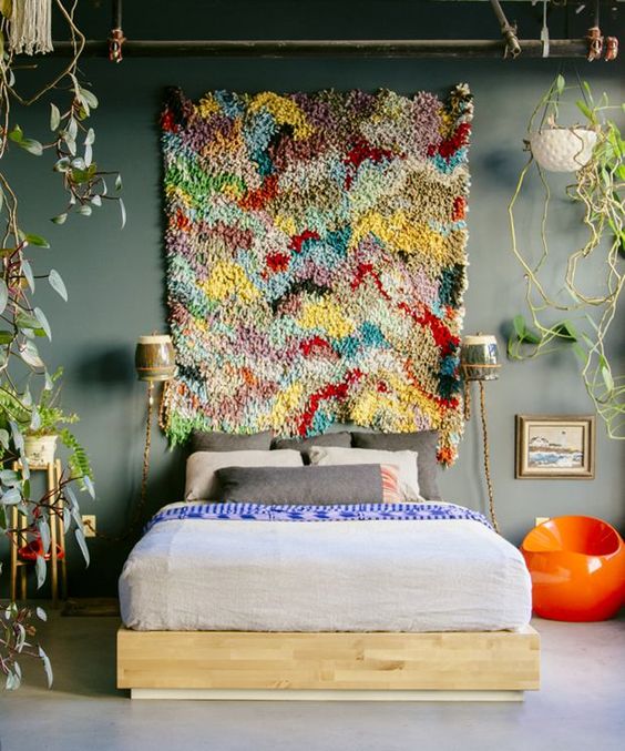 a grey bedroom with a bed, a colorful boho rug, potted greenery and an orange side table plus a lot of greenery around