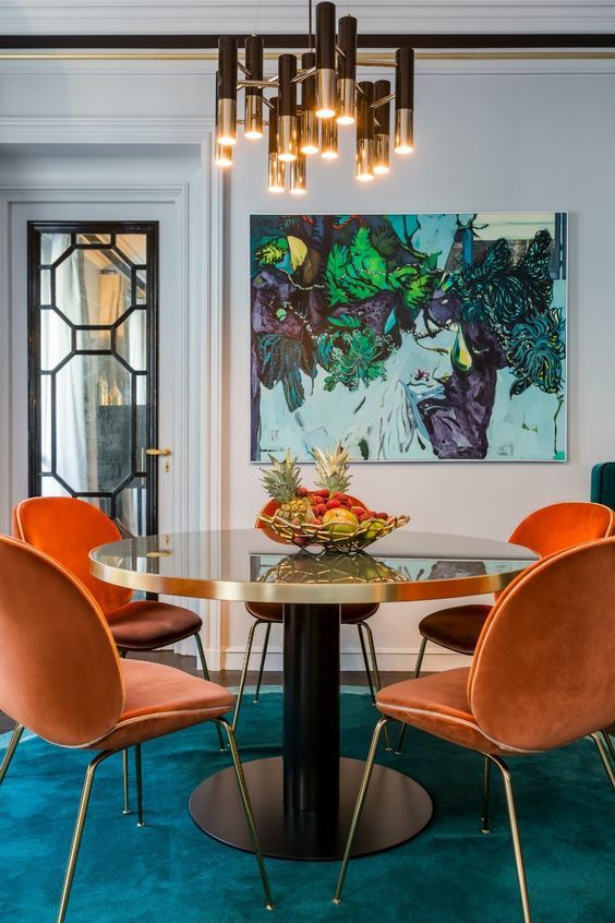 a jewel-tone dining room with a teal floor, orange chairs, a gold table, a bold artwork and a chic gold and black chandelier
