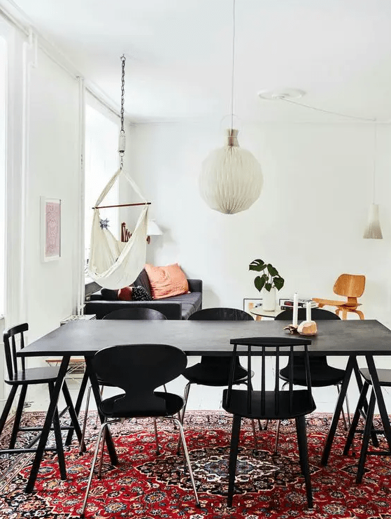 a living and dining room with a bold boho rug, a black table and mismatching chairs, a navy sofa with pillows, a chair and a table