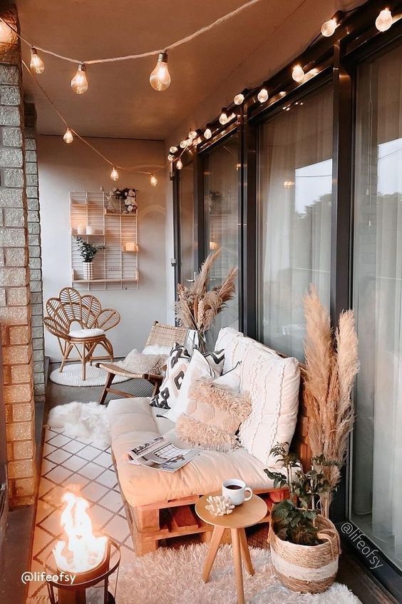 a lovely boho balcony with a pallet loveseat and pillows, a rattan and cane chair, pampas grass and greenery plus lights