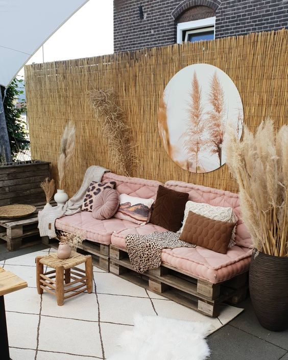 a lovely boho terrace with a pallet sofa in pink and lots of pillows, some stools and a table, pampas grass and a round mirror