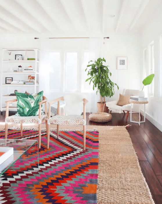 a lovely modern living room with layered rugs, a bold and a jute one, woven chairs, a white chair, a shelving unit and some greenery