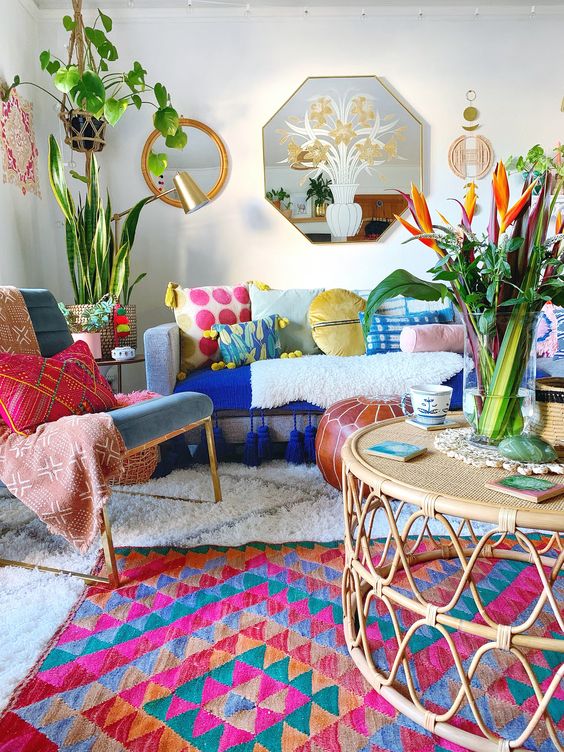 a maximalist living room with a blue sofa and colorful pillows, a bright rug, a blue chair and pillows, rattan furniture and potted greenery