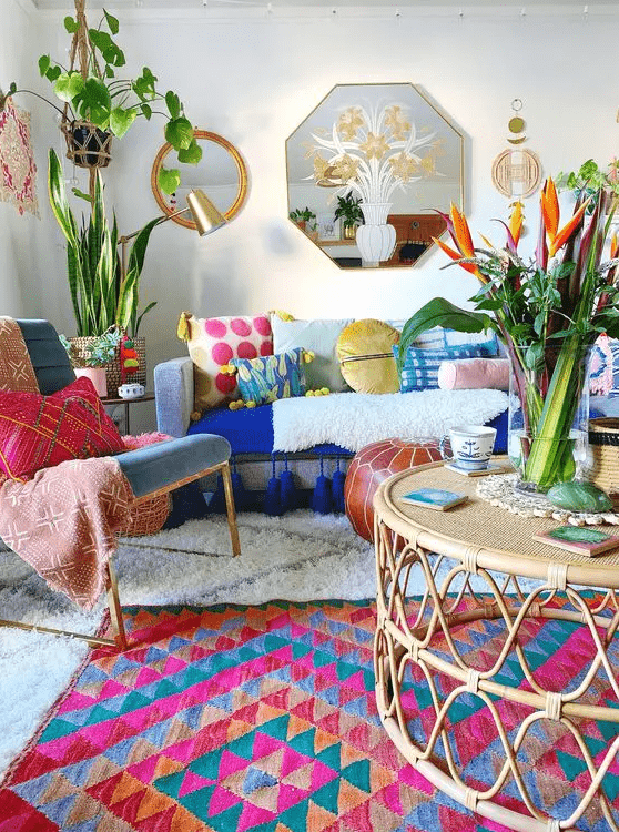 a maximalist living room with a blue sofa and colorful pillows, a bright rug, a blue chair and pillows, rattan furniture and potted greenery