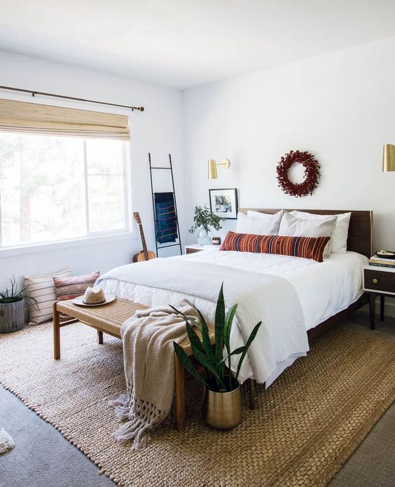 a modern bedroom with a bed and neutral bedding, a woven bench, a jute rug, some boho decor and potted greenery