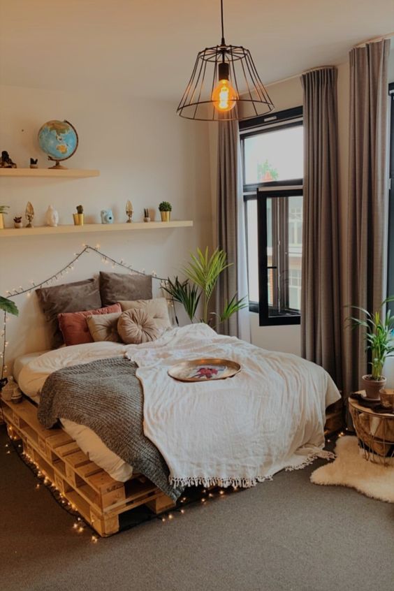 a modern boho bedroom with a pallet bed and neutral bedding, shelves with decor, lights, a pendant lamp and potted greenery