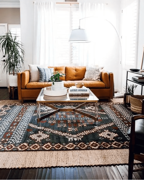 a modern boho living room an orange sofa and pillows, layered rugs including a jute one, a console table with decor, a floor lamp and greenery