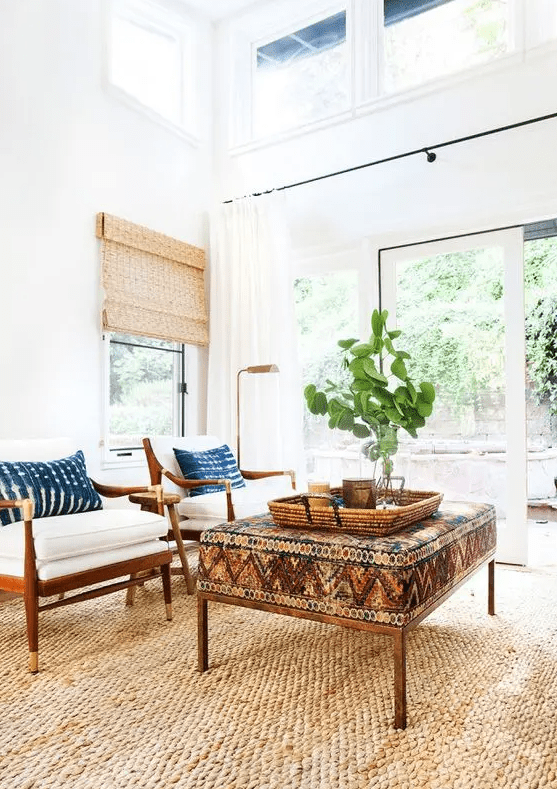 a modern boho living room with a jute rug, a bold boho ottoman, white chairs with coastal pillows, greenery and woven shades