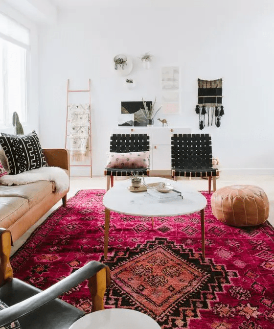 a modern boho living room with a pink boho rug, a tan leather couch, black chairs, a coffee table and a leather pouf and some wall decor