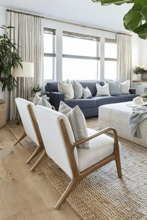 a modern coastal living room with a blue sofa, a neutral ottoman, white chairs, a jute rug, potted greenery and lamps