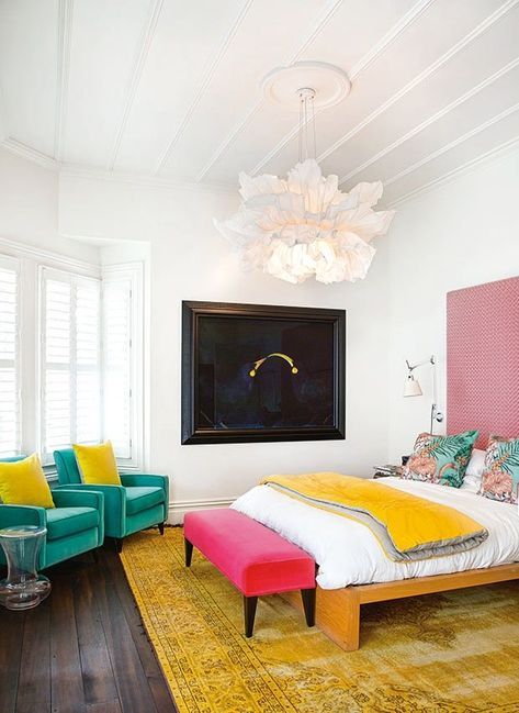a modern colorful bedroom with a pink accent wall, a yellow rug, a bed with bright bedding, a pink bench, emerald chairs