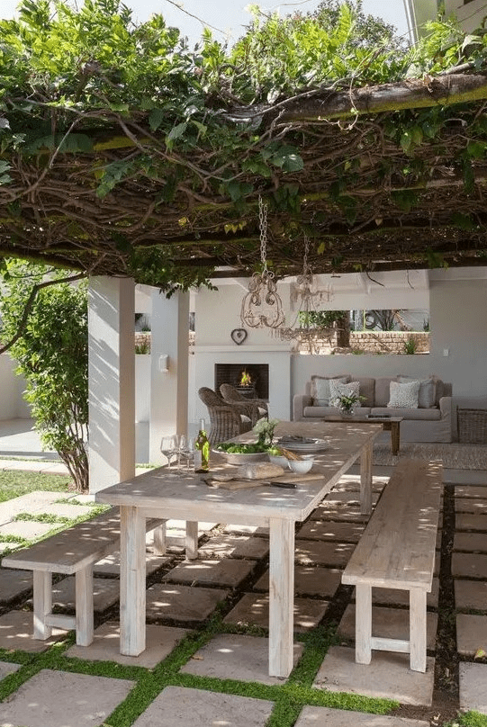 a modern country terrace with a living roof over the space, a whitewashed dining set with benches and an outdoor living room by the fireplace