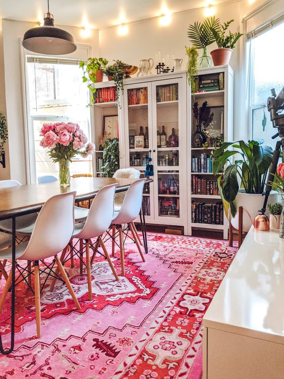a modern dining room done in neutrals, with a large bookcase, a table and white chairs, lots of potted greenery and a bold pink rug to add color